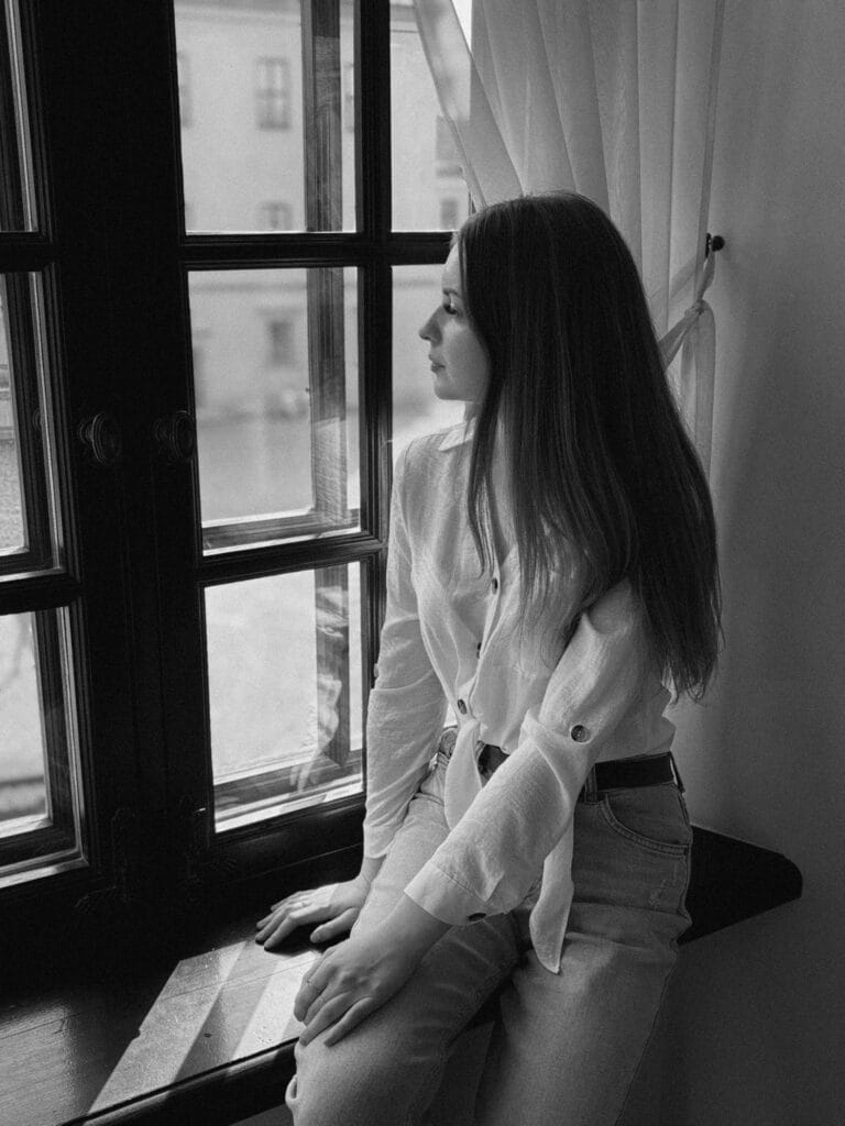Black and white portrait of a woman sitting by a window, lost in thought.